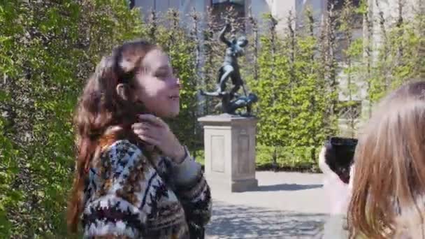 Dua perempuan muda berjalan di taman di antara tanaman dekoratif dan mengambil gambar dengan patung — Stok Video