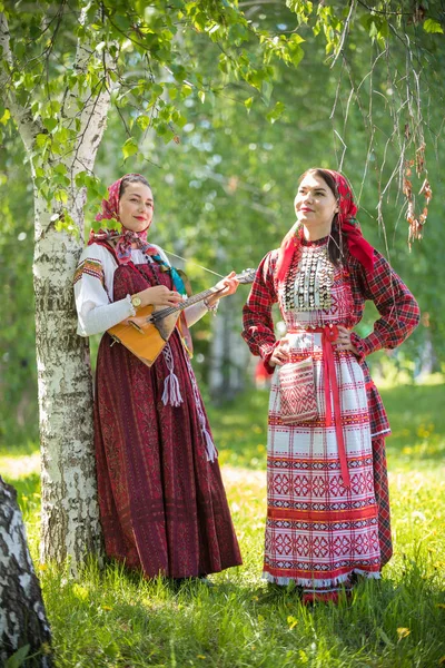 Duas mulheres sorridentes em roupas tradicionais russas em pé na floresta. Um deles segurando balalaika — Fotografia de Stock