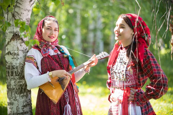 Two young woman in traditional russian clothes standing in the forest. One of them holding balalaika — Stock Photo, Image
