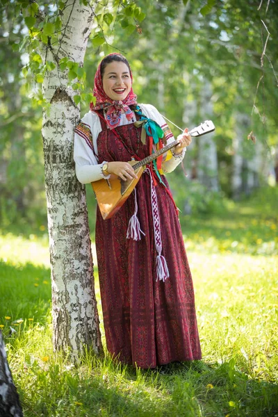 Smiling young woman in traditional russian clothes standing in the forest and playing balalaika — Stock Photo, Image