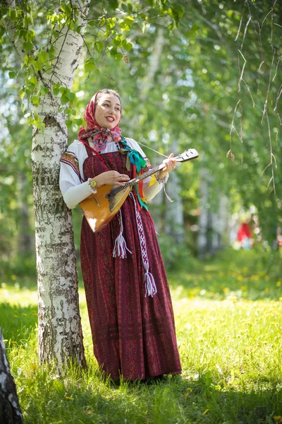 Молодая романтичная женщина в традиционной русской одежде стоит в лесу и держит балалайку — стоковое фото