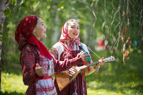 Dos mujeres jóvenes con ropa tradicional rusa cantando en el bosque. Uno de ellos tocando balalaika —  Fotos de Stock