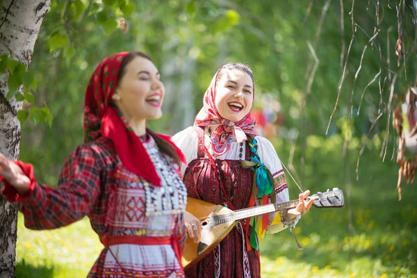 Two young attractive women in traditional russian clothes singing in the forest. One of them playing balalaika — Stock Photo, Image