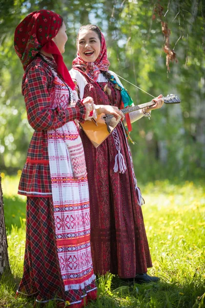 Two young woman in traditional russian clothes standing in the forest and look at each other. One of them playing balalaika and sing — Stock Photo, Image