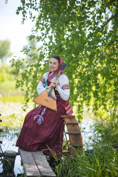 Молода усміхнена жінка в традиційному російському одязі сидить на маленькому мосту біля озера і тримає балаїку. — стокове фото