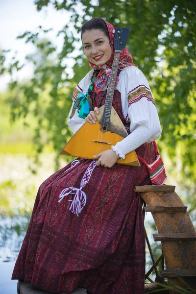 Geleneksel Rus kıyafetleri içinde genç romantik bir kadın gölün yanındaki küçük rıhtımda oturuyor ve kameraya poz veriyor. — Stok fotoğraf