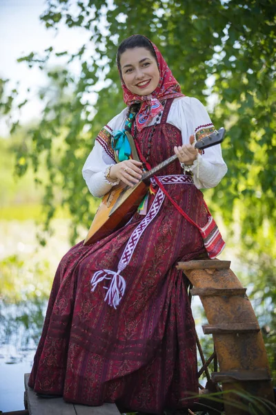 Mujer romántica joven en ropa tradicional rusa se sienta en el pequeño muelle cerca del lago, y reproducir música en balalaika —  Fotos de Stock