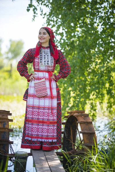 Mulher jovem em lenço e roupa russa tradicional está em um pequeno cais perto do lago, e mantém as mãos em uma cintura — Fotografia de Stock