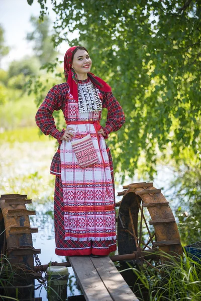 Jovem mulher em cachecol e roupas tradicionais russas fica em um pequeno cais perto do lago e mantém as mãos em uma cintura olhando para a esquerda — Fotografia de Stock