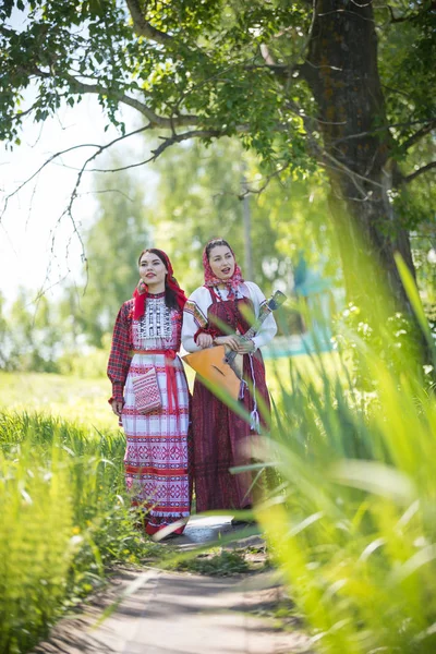 Dos mujeres jóvenes con ropa tradicional rusa caminando por el camino y hablando. Uno de ellos sosteniendo balalaika —  Fotos de Stock