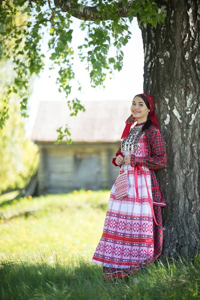 Молодая женщина в традиционной русской одежде стоит под деревом и позирует для фотографии — стоковое фото