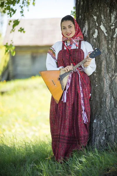 Joven mujer atractiva en ropa tradicional rusa de pie debajo de un árbol con la balalaika y posando para una foto —  Fotos de Stock