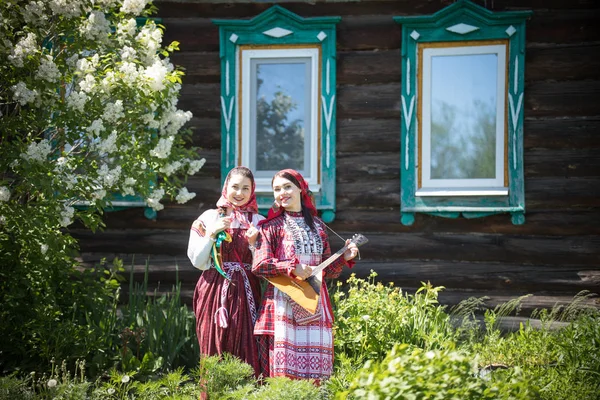 Две девушки в традиционной русской одежде стоят перед деревянным домом, одна из них играет в балалайку — стоковое фото
