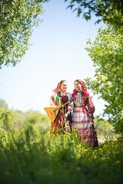 Δύο νεαρές γυναίκες σε παραδοσιακά ρωσικά ρούχα στέκονται στο χωράφι μεταξύ του πράσινου — Φωτογραφία Αρχείου