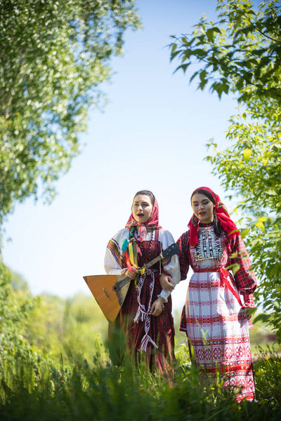 Two young women in traditional russian clothes stand in the field between the trees and singing