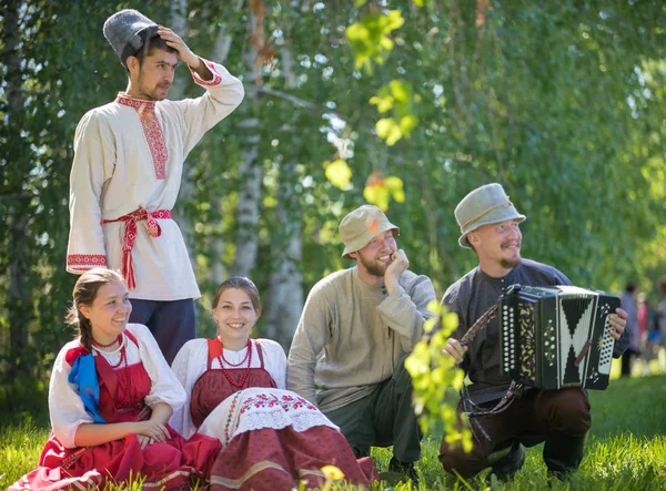 На лужайке сидят люди в традиционной русской одежде - один из них играет на аккордеоне — стоковое фото