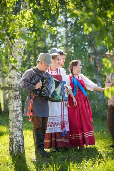 Lidé v tradičních ruských šatech stojí pod březným stromem a usmívají se-jeden z nich hraje na harmoniku — Stock fotografie