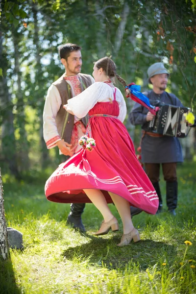 Мужчина и женщина в традиционной русской одежде кружатся в русском народном танце - рядом играет мужчина с аккордеоном — стоковое фото