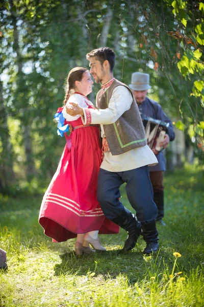 Άντρας και γυναίκα με παραδοσιακά Ρώσικα ρούχα πηδούν σε Ρώσους παραδοσιακούς χορούς κοντά σε έναν άντρα με ακορντεόν. — Φωτογραφία Αρχείου