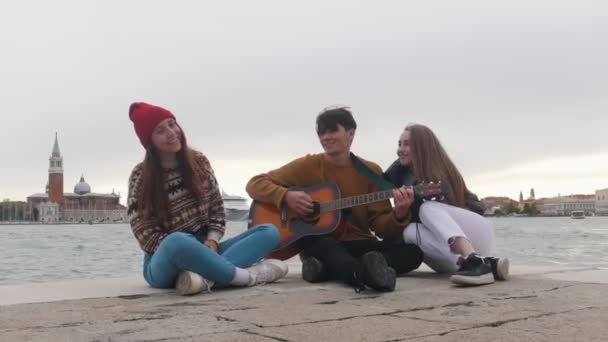 Un grupo de jóvenes amigos sentados en el borde del muelle y cantando canciones de la guitarra — Vídeos de Stock