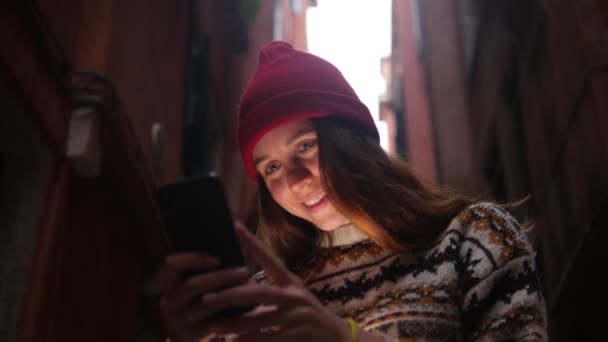 Una joven mujer sonriente de pie en la calle y mirando la pantalla del teléfono. Una luz ilumina su rostro — Vídeo de stock