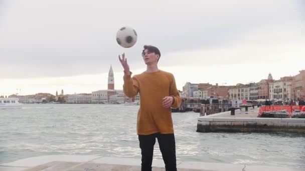 Un hombre jugando con una pelota de fútbol en el estrado sobre un fondo del mar. Girando la pelota en su dedo — Vídeos de Stock