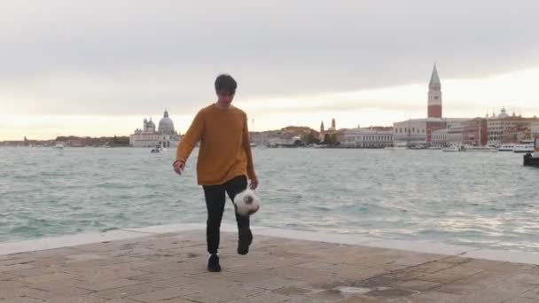Чоловік грає з футбольним м'ячем на підставці на фоні моря. Збиваючи м'яч ногами — стокове відео