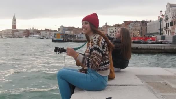 Молоді друзі сидять на краю набережної. Чоловік грає на гітарі — стокове відео