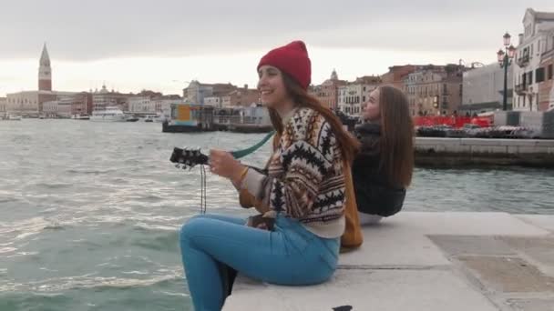 Jovens amigos sentados à beira do cais. Um homem a tocar guitarra — Vídeo de Stock