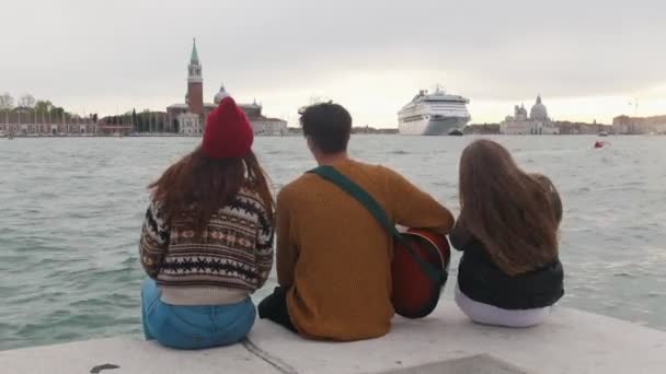 Jóvenes amigos sentados en el borde del muelle y disfrutando de la vista. Un hombre tocando la guitarra — Vídeo de stock