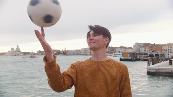 Um jovem que joga com uma bola de futebol no suporte em um fundo do mar. Girando a bola em seu dedo — Vídeo de Stock