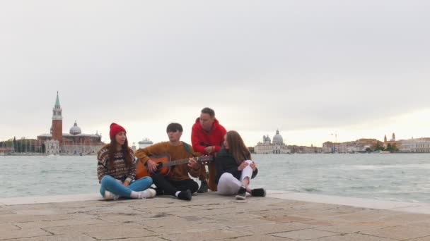 Um grupo de jovens amigos sentados à beira do cais e cantando canções. Um homem a tocar guitarra — Vídeo de Stock