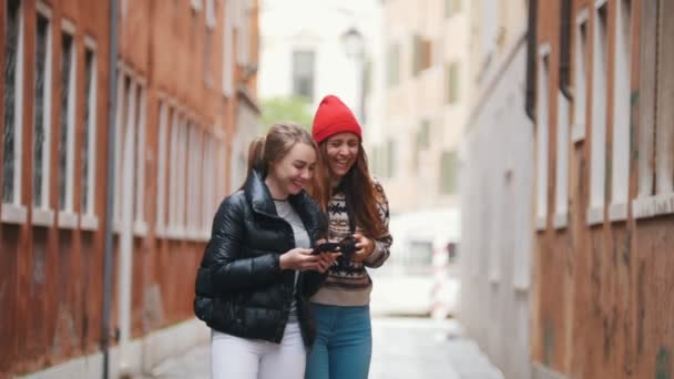 Zwei junge Frauen in warmen Kleidern gehen mit Handy und Kamera durch die engen Straßen. Frau zeigt ihr lustiges Foto am Telefon und sie lachen — Stockvideo