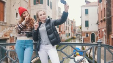 İtalya, Venedik. Küçük köprüde duran ve bir su kanalının arka planında selfie çeken iki genç kadın yolcu