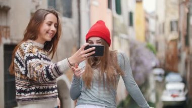 İtalya, Venedik. Küçük köprüde duran ve su kanalının arka planında selfie çeken iki genç kadın