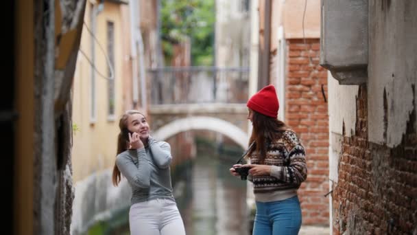 Zwei junge lachende Reisende in warmer Kleidung, die auf dem Hintergrund eines Wasserkanals stehen. eine Frau telefoniert, eine andere hält eine Kamera in der Hand — Stockvideo