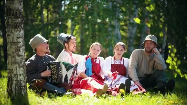 伝統的なロシアの服を着た人々は芝生の上に座り、アコーディオンで歌う — ストック動画