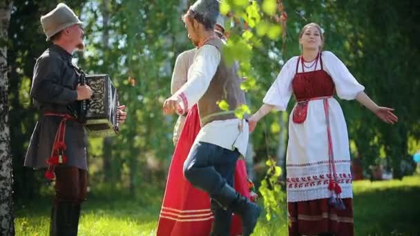 Människor i traditionella ryska kläder dansar på fältet håller händerna-en av dem spelar dragspels musik — Stockvideo