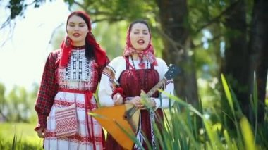 Geleneksel Rus kıyafetleri giyen iki genç kadın sahada yürüyüp şarkı söylüyor.