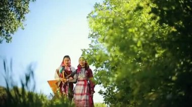 Geleneksel Rus kıyafetleri giyen iki genç kadın sahada yürüyüp şarkı söylüyor ve biri balalaika tutuyor..