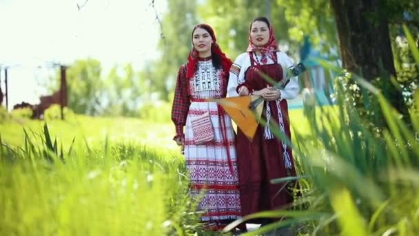Dos mujeres jóvenes con ropa tradicional rusa caminando por el campo y cantando - una de ellas sosteniendo balalaika — Vídeo de stock