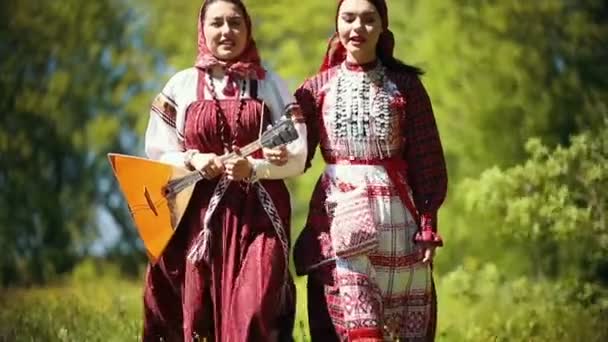 Zwei junge Frauen in traditioneller russischer Kleidung, die im Wald spazieren gehen und ein Lied singen - eine von ihnen mit Balalaika — Stockvideo