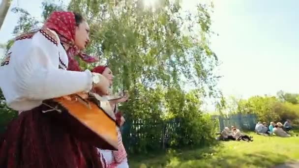 Oroszország, Nikolskoe falu, Köztársaság Tatarstan 25-05-2019: két fiatal nő a hagyományos orosz ruhák séta az úton a faluban, és énekel egy dalt játszik balalajka — Stock videók