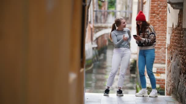 Zwei junge lachende Frauen, die auf dem Hintergrund eines Wasserkanals stehen und ins Telefon schauen — Stockvideo