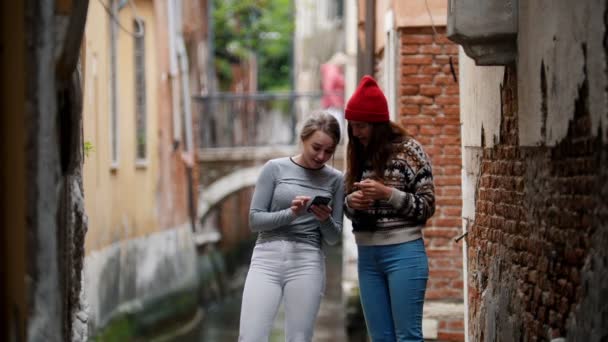 Zwei junge attraktive Frauen, die auf dem Hintergrund eines Wasserkanals stehen und Musik am Telefon hören. — Stockvideo