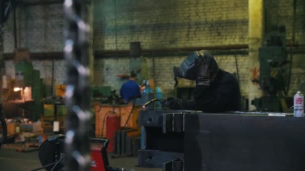 Usine de construction. Un homme dans un casque utilisant une machine à souder. Une chaîne suspendue au premier plan — Video