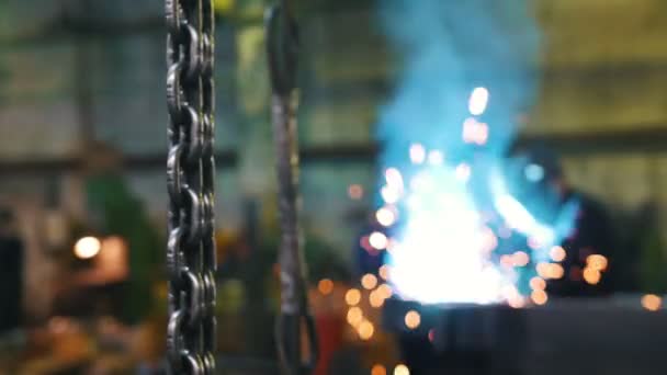 建筑厂。悬挂在前景上的金属链。一个男人焊接在背景与蓝色火花和光 — 图库视频影像