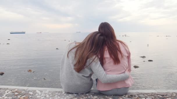 Dos mujeres jóvenes se abrazan sentadas en las escaleras cerca del mar mirando el agua — Vídeos de Stock