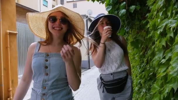 Dos jóvenes mujeres felices caminando en panamas en las calles a lo largo de un frondoso muro — Vídeo de stock