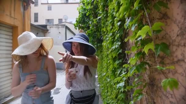 Two young women walking in panamas on the streets along a leafy wall. One of them drinking coffee from a cup — Stock Video